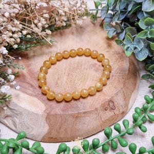 May include: A golden-yellow beaded bracelet rests on a wooden surface, surrounded by greenery and dried flowers. The bracelet is made of small, round beads, showcasing a warm, amber hue. The natural setting enhances the bracelet's earthy tones.