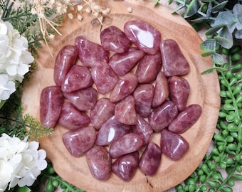 May include: A collection of polished, tumbled rose quartz stones in varying shades of pink and red. The stones are arranged on a round, wooden plate, surrounded by greenery and white flowers. The stones have a smooth, glossy finish.