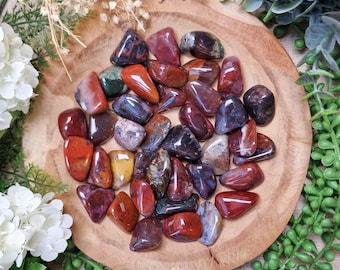 May include: A collection of polished gemstones in shades of red, brown, and purple, displayed on a wooden plate. The stones vary in shape and size, with some displaying unique patterns and textures. The arrangement is surrounded by greenery and white flowers.