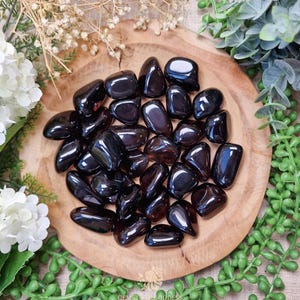 May include: A selection of glossy, black gemstones displayed on a circular wooden platter. The stones are of varying sizes and shapes. The wooden platter is surrounded by green foliage and white flowers, creating a natural display.