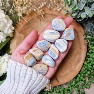 May include: A handful of tumbled stones in shades of blue, cream, and gold. The stones have a smooth, polished appearance and are held in an open hand. The background includes greenery and a wooden surface. The logo "GEMSTONE GODDESS" is visible at the bottom.