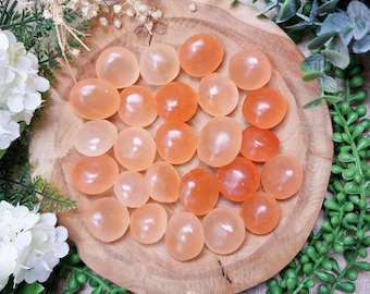 May include: A collection of smooth, orange-toned gemstones arranged on a round, wooden plate. The stones vary in color from light peach to a deeper orange. The arrangement is surrounded by greenery and white flowers, creating a natural aesthetic.