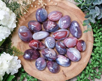 May include: A collection of polished agate stones in shades of purple, red, and white, displayed on a wooden plate. The stones vary in size and shape, with visible banding patterns. The image is surrounded by white flowers and green foliage.