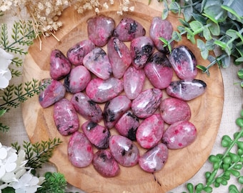 May include: A collection of polished, oval-shaped gemstones in shades of pink and black, arranged on a wooden plate. The stones vary in color intensity, with some displaying darker pink and black inclusions. The arrangement is surrounded by greenery and dried flowers.