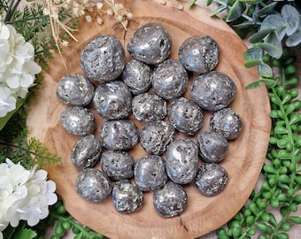 May include: A collection of pyrite stones, also known as fool's gold, arranged on a wooden plate. The stones have a metallic, silver-gray color and a rough, textured surface. The plate is surrounded by greenery and white flowers, creating a natural aesthetic.