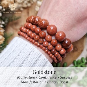 Può includere: Diversi braccialetti di perline di pietra dorata di varie dimensioni sono impilati su un polso. Le perline hanno un aspetto marrone-rossastro scintillante. L'immagine include il testo "Goldstone" e le parole "Motivazione, Fiducia, Successo, Manifestazione, Aumento di energia."