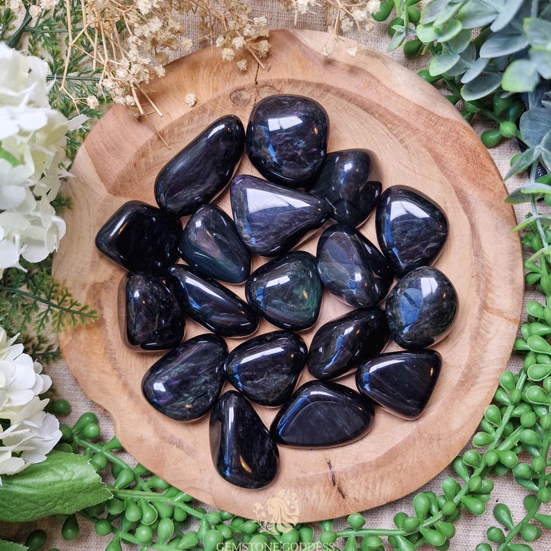 May include: A collection of tumbled black obsidian stones with iridescent blue and green flashes. The stones are arranged in a wooden bowl.
