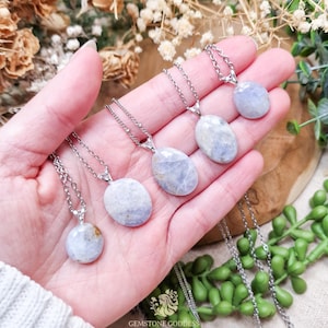 May include: Five gemstone pendant necklaces with oval and round light blue stones on silver chains. The necklaces are displayed in a hand, showcasing the variety of shapes and sizes. The background includes greenery and dried flowers.
