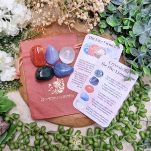 May include: A selection of gemstones in shades of red, blue, and clear, presented with information cards. The gemstones are in a pink drawstring bag with the text "GEMSTONE GODDESS". The cards detail the five elements and their corresponding crystals.