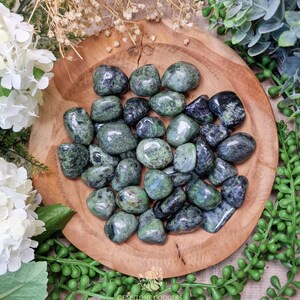 May include: A collection of green and black tumbled stones in a wooden bowl. The stones are smooth and polished, and they have a variety of patterns and textures.