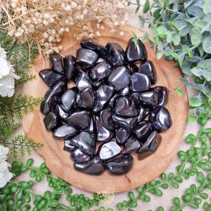 Op de afbeelding: Een verzameling gepolijste, donkergrijze hematietstenen, gepresenteerd in een ronde houten kom. De stenen hebben een reflecterende, metallic glans. De opstelling is omgeven door groen en gedroogde bloemen.