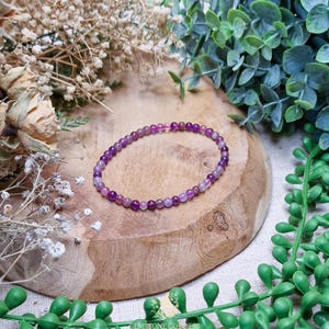 May include: A bracelet composed of small, round, purple gemstones. The bracelet is presented on a wooden surface, with greenery and dried flowers surrounding it. The gemstones display varying shades of purple, creating a subtle gradient effect.