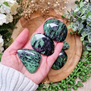 Könnte beinhalten: Fünf glatte, polierte Zoisit-Steine in Grün, Schwarz und Rosa. Die Steine liegen in der Handfläche.