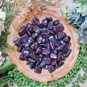 May include: A bowl filled with tumbled, polished, dark purple and black hematite stones. The stones have a metallic sheen and are smooth to the touch.