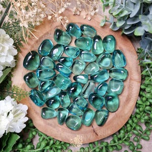 Puede incluir: Una colección de piedras preciosas pulidas de color verde azulado dispuestas sobre un plato de madera redondo. Las piedras tienen una apariencia suave y redondeada y varían en tamaño y forma. El plato está rodeado de vegetación y flores blancas. El texto "GEMSTONE GODDESS" es visible.