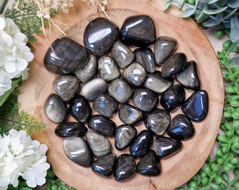 May include: A collection of polished silver sheen obsidian stones with a smooth, reflective surface and dark grey to black coloration. The stones are arranged in a cluster on a round wooden surface, showcasing natural metallic lustre and variations.