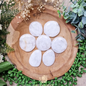 Peut inclure: Sept pierres ovales lisses et polies de couleur blanche disposées en forme de fleur sur une surface en bois.