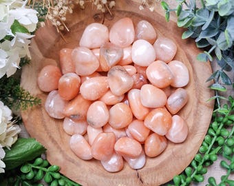 May include: A wooden bowl filled with smooth, polished peach-colored gemstones. The stones vary in size and shape, with some displaying translucent qualities. The bowl is surrounded by greenery and white flowers, creating a natural aesthetic.