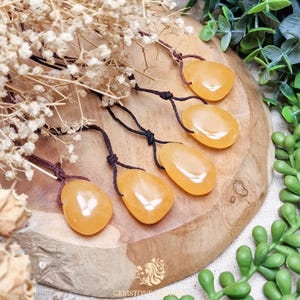 May include: Four polished, teardrop-shaped orange gemstone pendants, each suspended from a thin, dark brown adjustable cord. The smooth, translucent stones exhibit a warm, vibrant orange hue and are arranged in a cluster on a wooden surface.