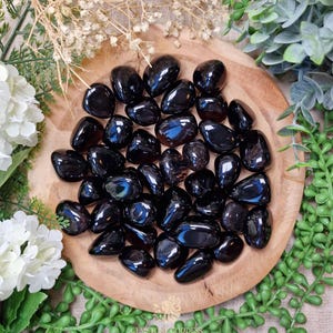 Puede incluir: Un conjunto de piedras de obsidiana negra pulidas, dispuestas sobre una bandeja de madera redonda. Las piedras tienen una superficie brillante y reflectante. El arreglo está rodeado de vegetación y flores blancas, creando una estética natural.