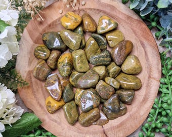 May include: A wooden bowl filled with polished gemstones. The stones are a mix of olive green, brown, and yellow, with a smooth, rounded shape. The gemstones are arranged in a circular pattern. The bowl is surrounded by greenery and white flowers.