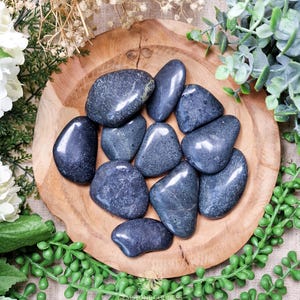 Pode incluir: Uma coleção de pedras lisas de cor cinza escuro, provavelmente obsidiana, dispostas em uma tigela de madeira. As pedras têm um brilho ligeiramente iridescente e são uma escolha popular para cura de cristal e meditação.