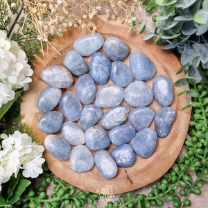 Può includere: Una collezione di pietre burattate lisce, blu-grigie, disposte su un piatto di legno, circondate da verde e fiori bianchi. Le pietre variano per dimensioni e forma, con un aspetto lucido e brillante. L'estetica generale è naturale e serena.