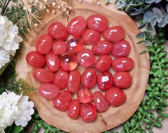 May include: A collection of polished, red-toned gemstones arranged on a wooden plate, surrounded by greenery and white flowers. The gemstones vary in size and shape, with a glossy, translucent appearance. The text "GEMSTONE GODDESS" is visible at the bottom.