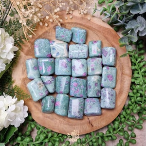 Può includere: Una collezione di pietre preziose burattate, principalmente turchesi con inclusioni rosa e bianche, disposte su un piatto di legno. Le pietre sono rettangolari e lisce, con un aspetto lucido. La disposizione è circondata da verde e fiori bianchi.