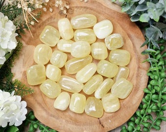 May include: A collection of polished yellow calcite stones displayed on a wooden bowl, surrounded by greenery and white flowers. The stones vary in size and shape, with a smooth, rounded appearance.