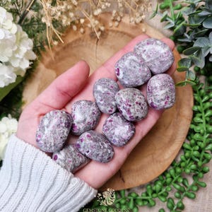 Könnte beinhalten: Eine Sammlung von getrommelten Steinen, hauptsächlich grau mit lebendigen rosa und roten Einschlüssen, wird in einer Hand gehalten. Die Steine haben ein glattes, poliertes Aussehen. Der Hintergrund umfasst eine Holzoberfläche und Grün.