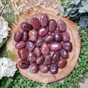 May include: A selection of polished, oval gemstones in shades of red and maroon. The stones are displayed on a round wooden plate, with greenery and white flowers around them. The gemstones have a glossy finish and are of various sizes.