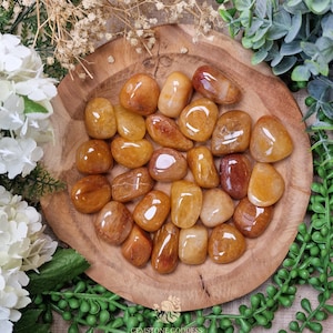 May include: A selection of polished, golden-yellow gemstones displayed on a round wooden tray. The stones vary in shade, with some displaying a deeper amber hue. The arrangement is surrounded by greenery and white flowers, creating a natural aesthetic.