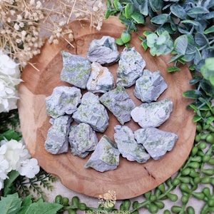 May include: A collection of rough, gray-green gemstones displayed on a wooden, circular tray. The stones have a mottled appearance with patches of green and gray. The arrangement is surrounded by greenery and white flowers, creating a natural aesthetic.