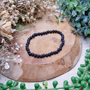 Può includere: Un braccialetto di perline di pietra lavica nera. Il braccialetto è composto da piccole perline rotonde e scure infilate insieme. Il braccialetto è esposto su una superficie di legno con fiori secchi e verde sullo sfondo.
