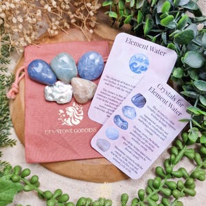 May include: A collection of polished gemstones in shades of blue, gray, and beige, displayed with informational cards. The stones are arranged on a pink drawstring bag, with text that reads "GEMSTONE GODDESS". The cards describe the properties of the stones.