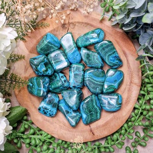 May include: A collection of tumbled azurite malachite stones in various shades of blue and green. The stones are arranged in a wooden bowl.