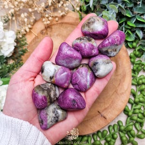 Può includere: Un gruppo di pietre preziose di rubino lucide, che mostrano una vibrante tonalità rosa-rossa con inclusioni grigie e bianche. Le pietre sono lisce e arrotondate, su una superficie di legno. L'immagine include il testo "GEMSTONE GODDESS".