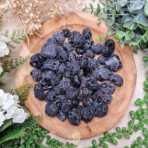 May include: A collection of polished black tourmaline stones displayed on a round wooden plate. The stones vary in size and shape, with a glossy, dark appearance. The plate is surrounded by greenery and white flowers.