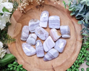 May include: A collection of tumbled white and lavender banded agate stones. The stones are smooth and polished, with a variety of shapes and sizes. The stones are arranged in a wooden bowl.