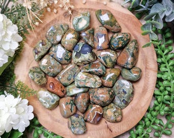 May include: A collection of polished gemstones in shades of green, brown, and cream, arranged on a wooden plate. The stones vary in size and shape, with a glossy finish. The arrangement is surrounded by greenery and white flowers.