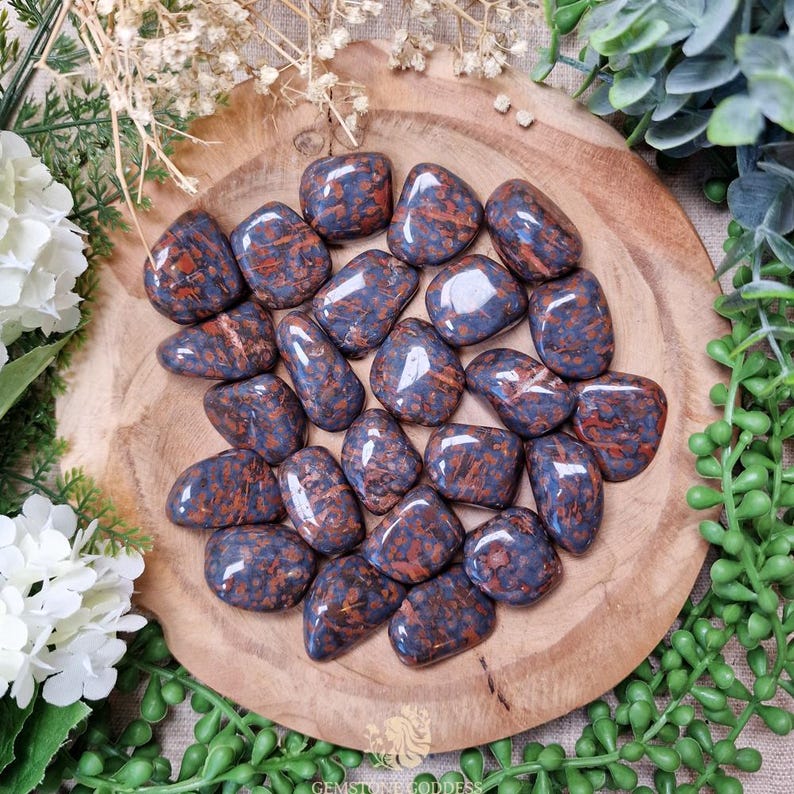 May include: A collection of polished gemstones in shades of blue and reddish-brown, arranged on a wooden surface. The stones have a smooth, rounded shape and a glossy finish. The image is surrounded by greenery and white flowers.
