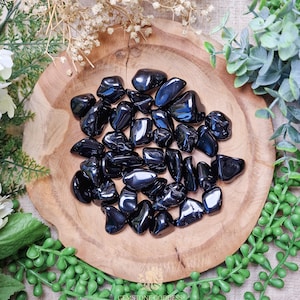Könnte beinhalten: Eine Sammlung von geschliffenen schwarzen Obsidian-Steinen in einer Holzschale. Die Steine sind glatt und poliert und haben eine glänzende Oberfläche.