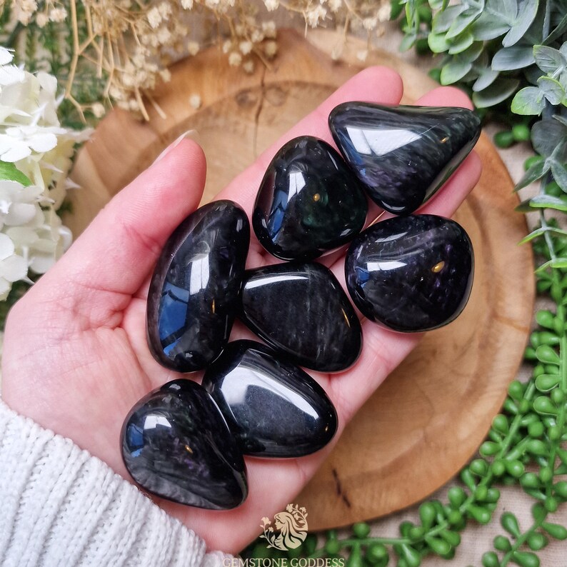 May include: A collection of seven black obsidian tumbled stones with iridescent shimmer. The stones are smooth and polished, and they are arranged in a hand.
