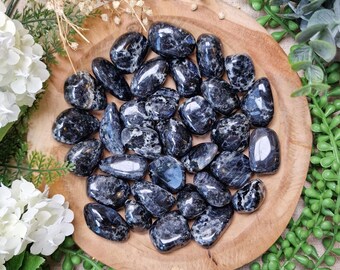 May include: A collection of polished blue-black stones with white marbling, arranged on a round wooden tray. The stones vary in size and shape, with a glossy finish. The tray is surrounded by white flowers and green foliage.