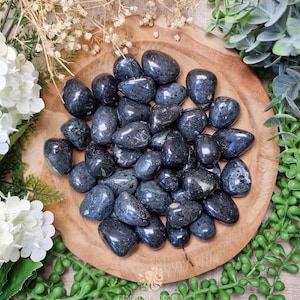 May include: A collection of polished, dark blue gemstones with iridescent flashes, displayed on a round wooden tray. The stones vary in size and shape, with a smooth, glossy finish. The arrangement is surrounded by white flowers and green foliage.