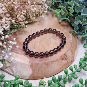 Puede incluir: Una pulsera de cuentas marrón oscuro hecha de piedras redondas y pulidas. La pulsera se exhibe sobre una superficie de madera, rodeada de vegetación y flores secas. Las cuentas tienen aproximadamente 0,8 cm de diámetro.