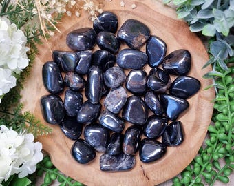 May include: A collection of polished, dark gray gemstones, possibly hematite, arranged on a round wooden surface. The stones have a smooth, reflective surface. The image includes the text "GEMSTONE GODDESS" at the bottom. Surrounding the stones are white flowers and green foliage.
