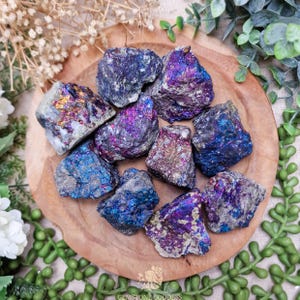 May include: A selection of iridescent pyrite stones, showing vivid shades of blue, purple, and gold. The rough gemstones are arranged on a round wooden tray, surrounded by greenery and white flowers. The stones vary in size and shape.