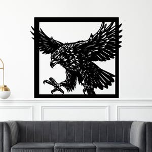 May include: Black and white wall art featuring a detailed illustration of an eagle in mid-flight. The eagle is enclosed within a black square frame, with its wings spread wide and talons extended. The artwork is mounted on a white wall.